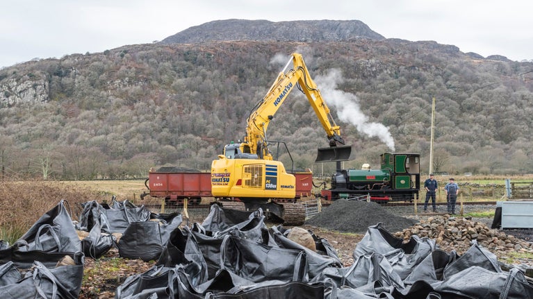 A yellow digger in front of a steam engine, with dozens of black sacks at the front of the picture waiting to be loaded